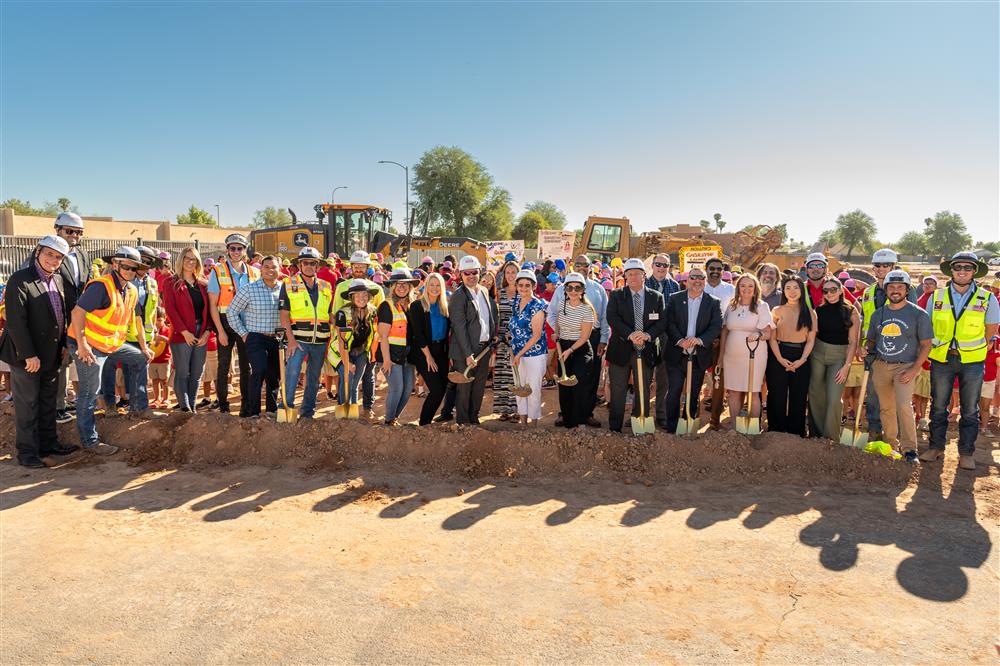 People dressed in hard hats and safety vests use shovels to dig into the ground